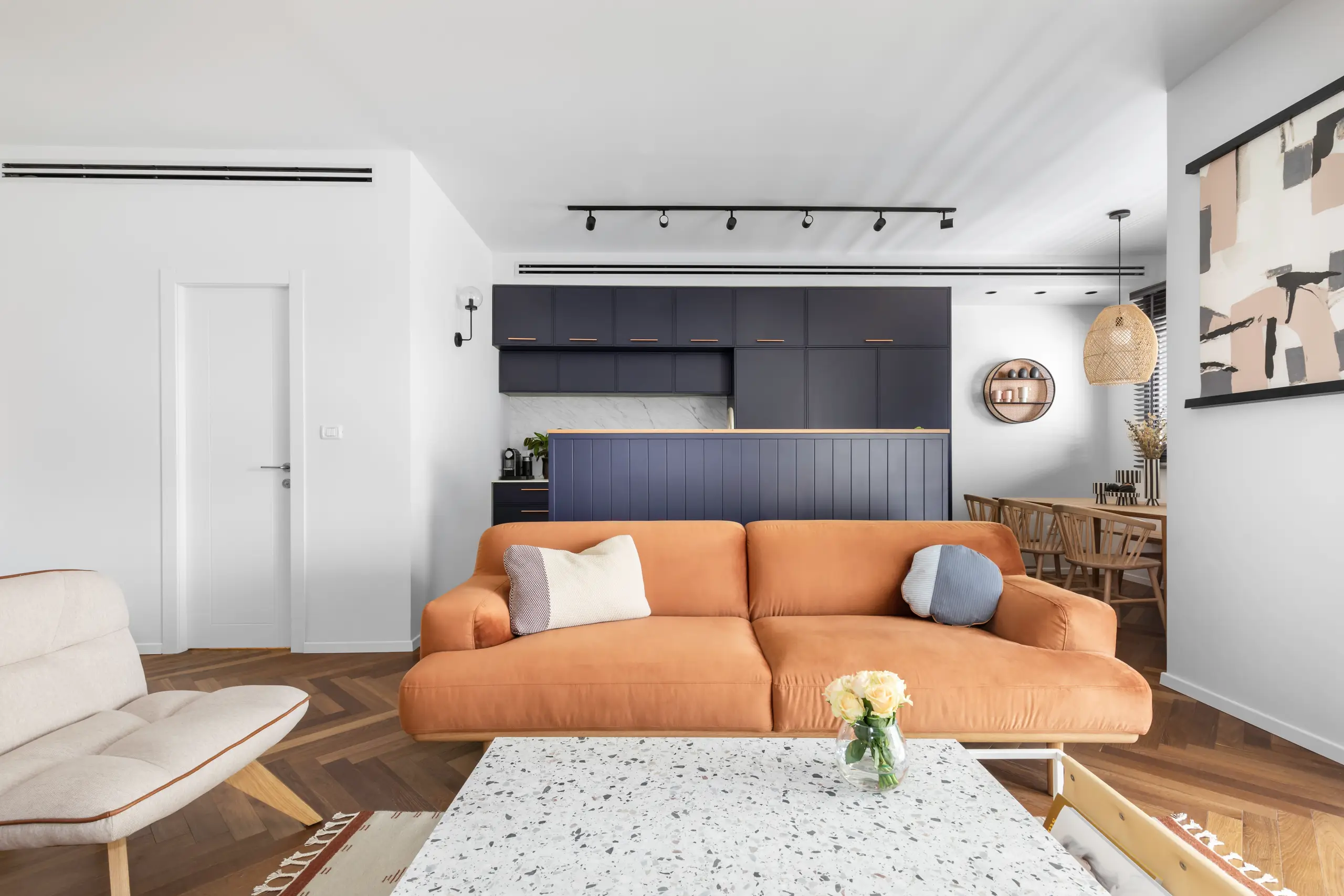 Orange sofa on a herringbone oak floor with a navy kitchen block beyond